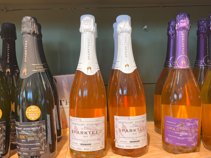 Bouteilles de thé pétillant sur une étagère en métal, avec des étiquettes de différentes couleurs, et des boîtes en carton en dessous.