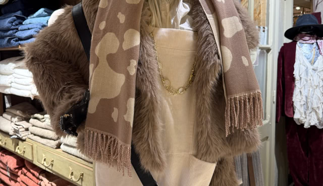 Mannequin vêtu d'un manteau en fourrure synthétique marron, écharpe beige à motifs, chapeau beige, pantalon clair, et sac en bandoulière noir, dans un magasin de vêtements.
