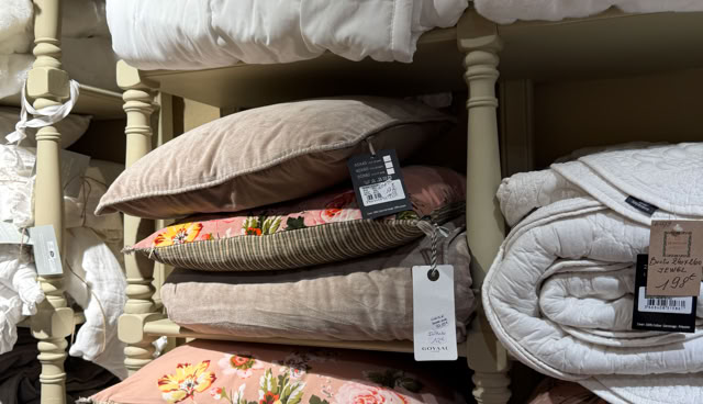 Oreillers et couvertures empilés sur des étagères, certains avec des motifs floraux et des étiquettes attachées.