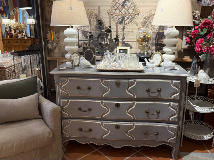 Un fauteuil beige à gauche, une commode grise avec trois tiroirs au centre, deux lampes avec abat-jour blancs sur la commode, des décorations et des fleurs rouges à droite.