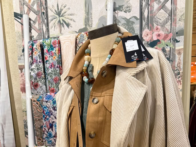 Mannequin portant un manteau beige et une chemise marron, avec un sac à main perforé marron en bandoulière, entouré de sacs et d'écharpes colorées.