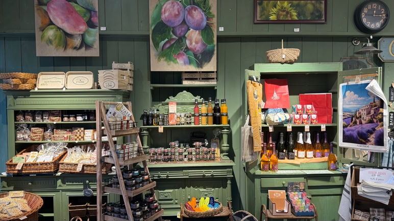 Intérieur d'une boutique avec des étagères vertes contenant des produits divers, des bouteilles, des paniers, et des cartes postales. Des peintures de fruits sont accrochées au mur.