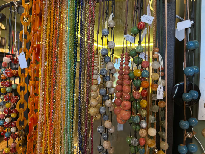 Colliers colorés suspendus, composés de perles de différentes tailles et couleurs, avec des étiquettes blanches attachées.