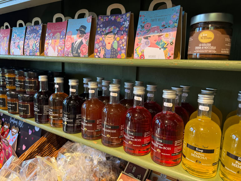Intérieur d'une boutique avec des étagères vertes contenant des produits divers, des bouteilles, des paniers, et des cartes postales. Des peintures de fruits sont accrochées au mur.
